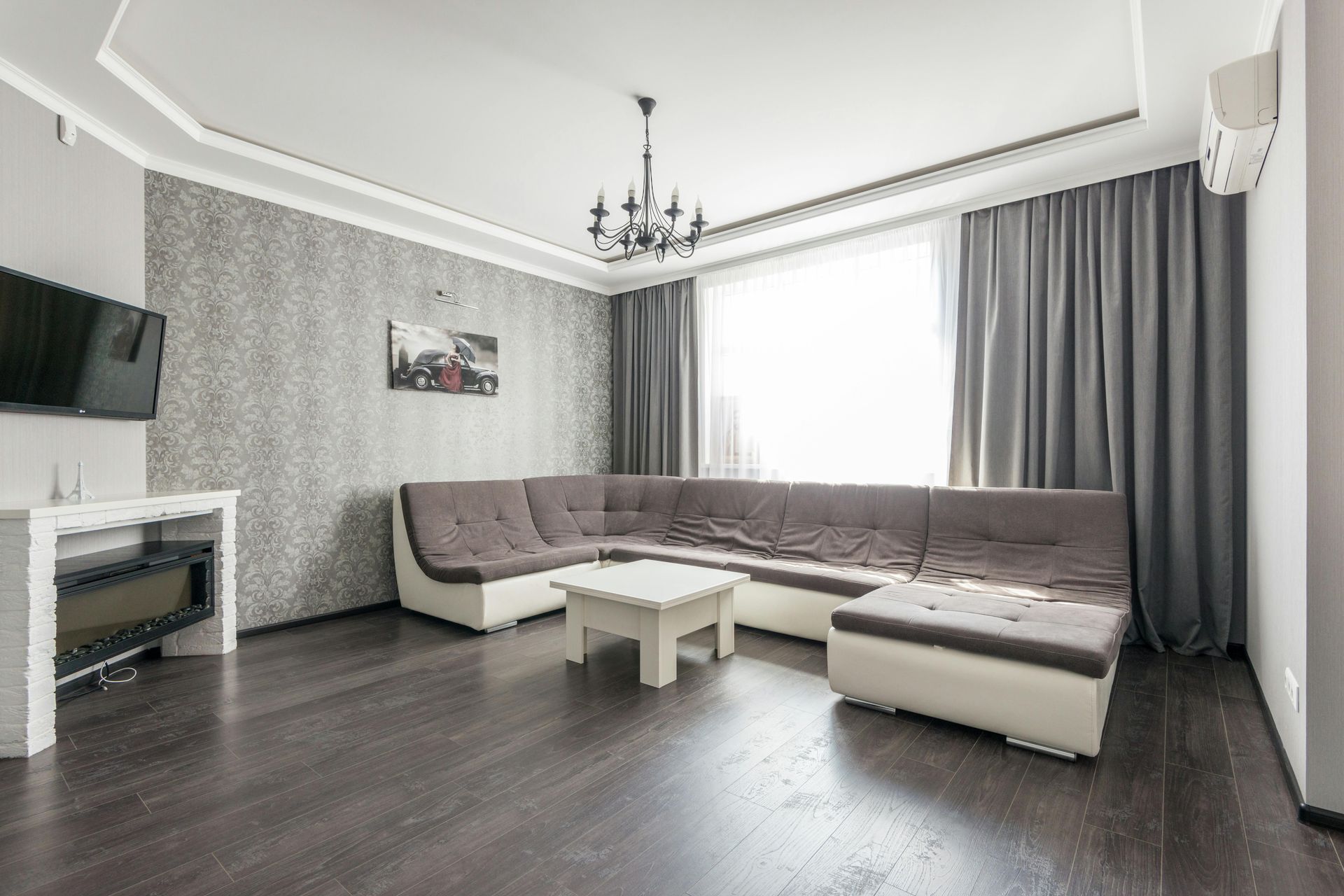 Bright living room with gray sectional sofa, white coffee table, TV, and curtains on dark flooring