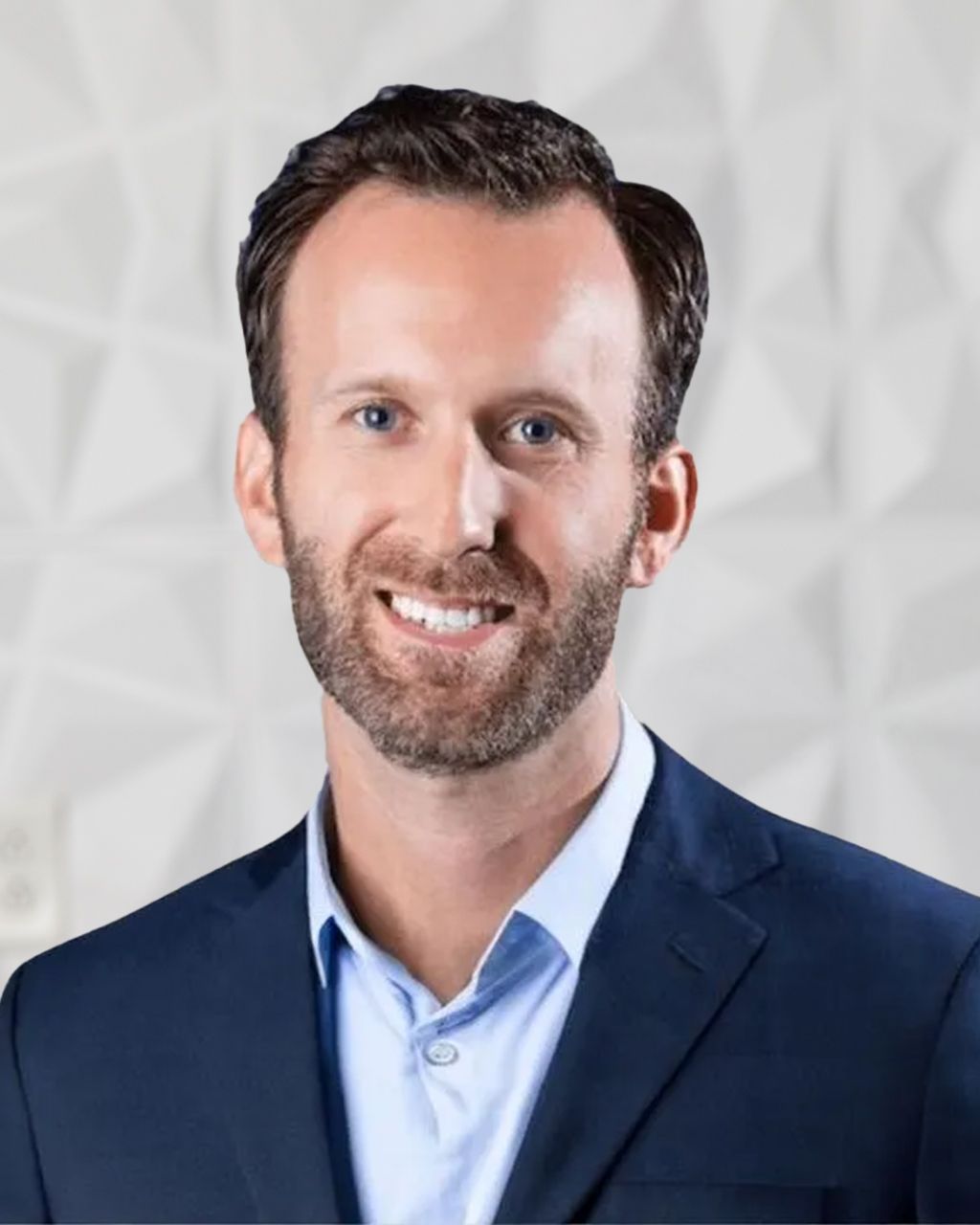 Man with brown hair and beard smiles, wearing a blue shirt and navy blazer against a textured white backdrop.