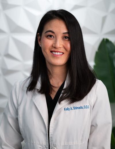 Woman in a white lab coat smiles. She has dark hair, and stands in front of a white patterned wall.