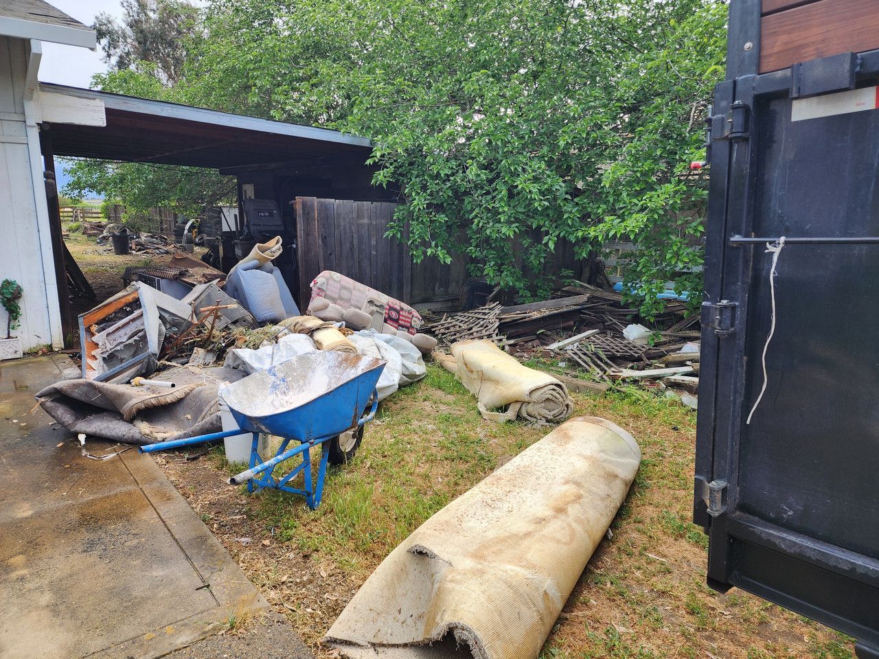 A wheelbarrow is sitting in the grass next to a dumpster in a backyard.