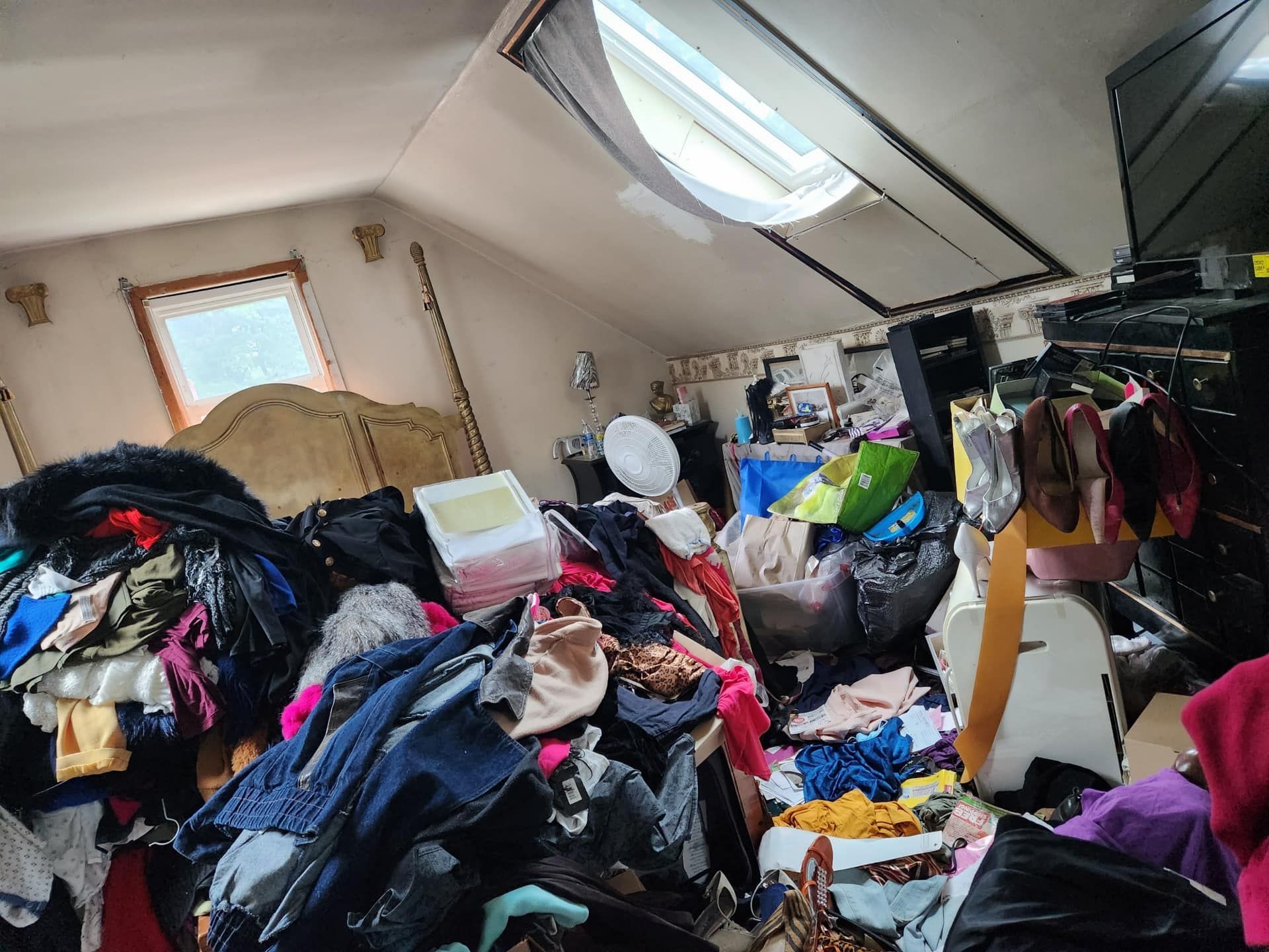 A bedroom overwhelmed by clutter, with a sea of clothes covering 90% of the visible floor and furniture, including a dresser and bed, highlighting a hoarding scenario.
