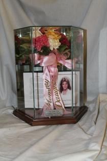 Floral memorial display in a glass case with a pink ribbon and a framed portrait on a wooden base