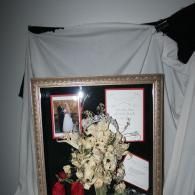 Framed memorial display with white flowers, photos, and notes against a draped white backdrop