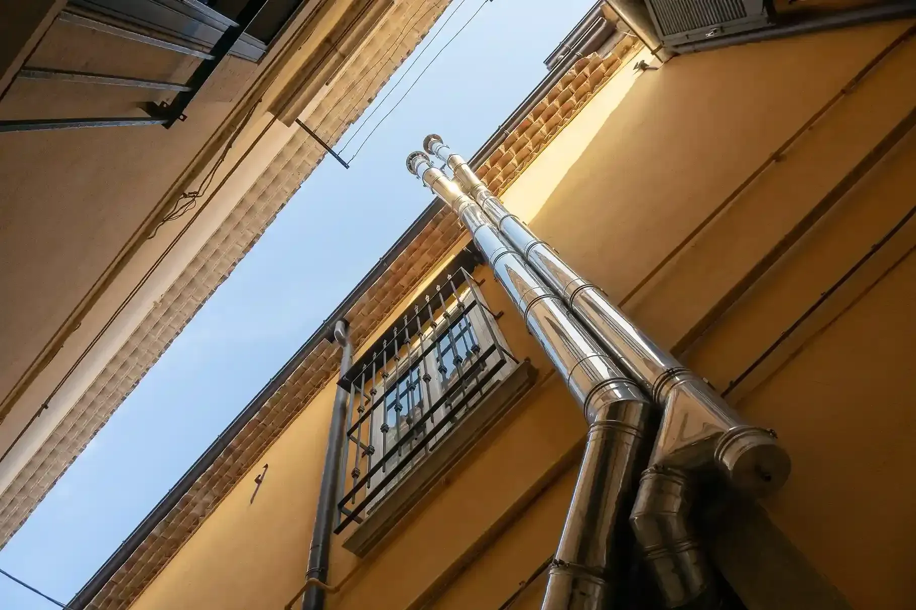 Low-angle view of building exteriors and silver metal pipes extending upwards against a blue sky.
