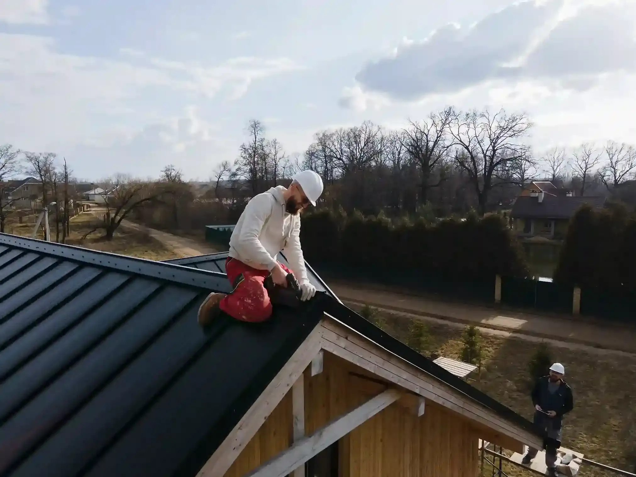 Person in hardhat works on black metal roof, another person below; sunny day.