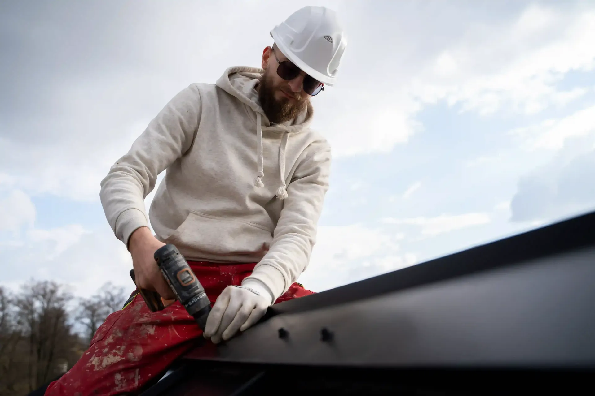 A person in a hard hat and gloves is applying sealant to a dark roof under a cloudy sky.