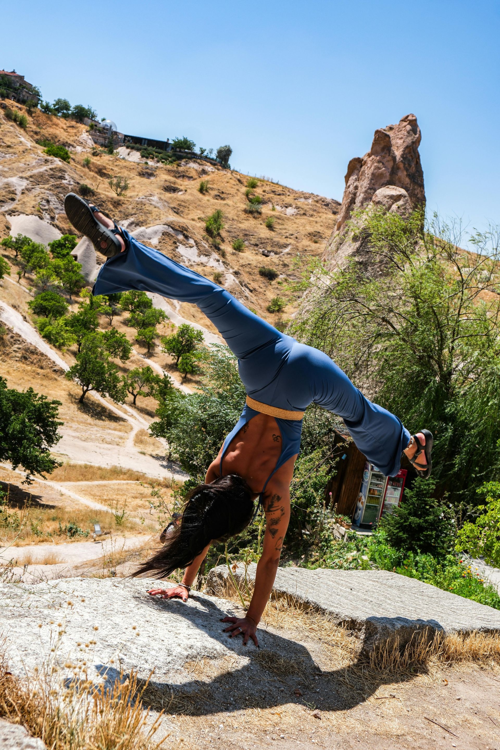 Woman in blue doing a handstand on a rock in a sunny, dry, mountainous landscape.