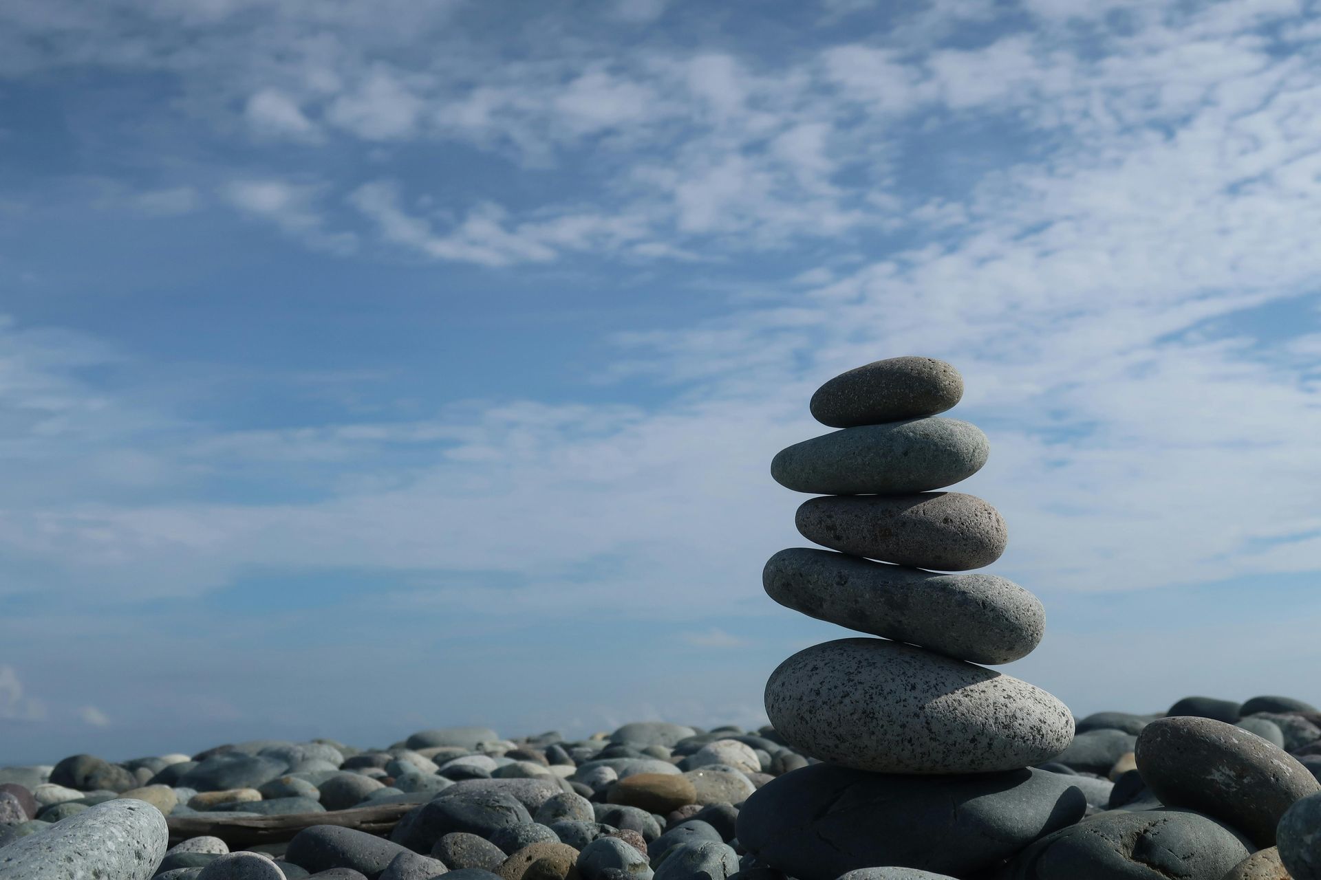 Stack of gray stones balanced on a pebble beach with a blue sky and clouds in the background.