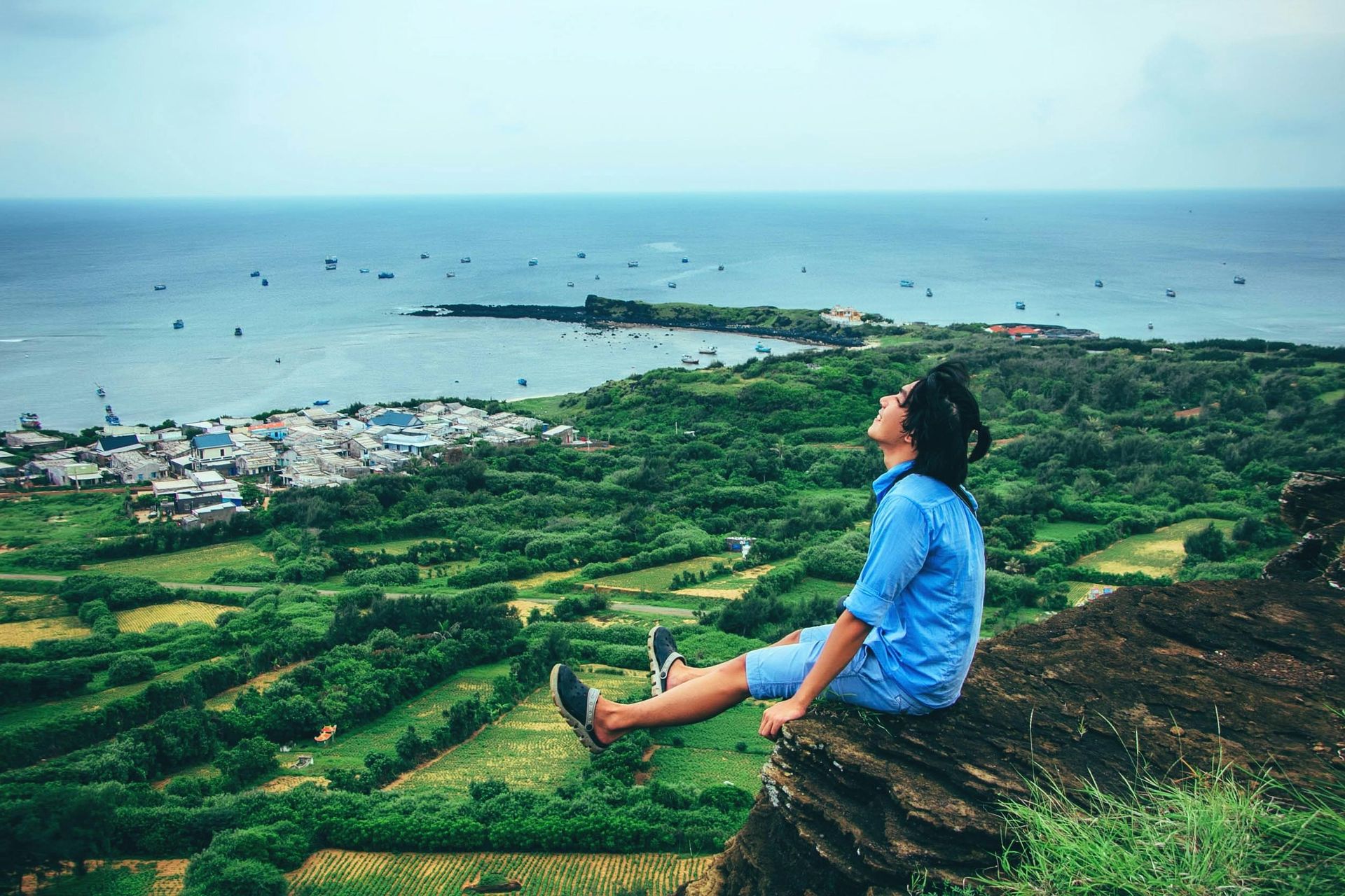 Person sits on a cliff overlooking a lush green landscape and the ocean.