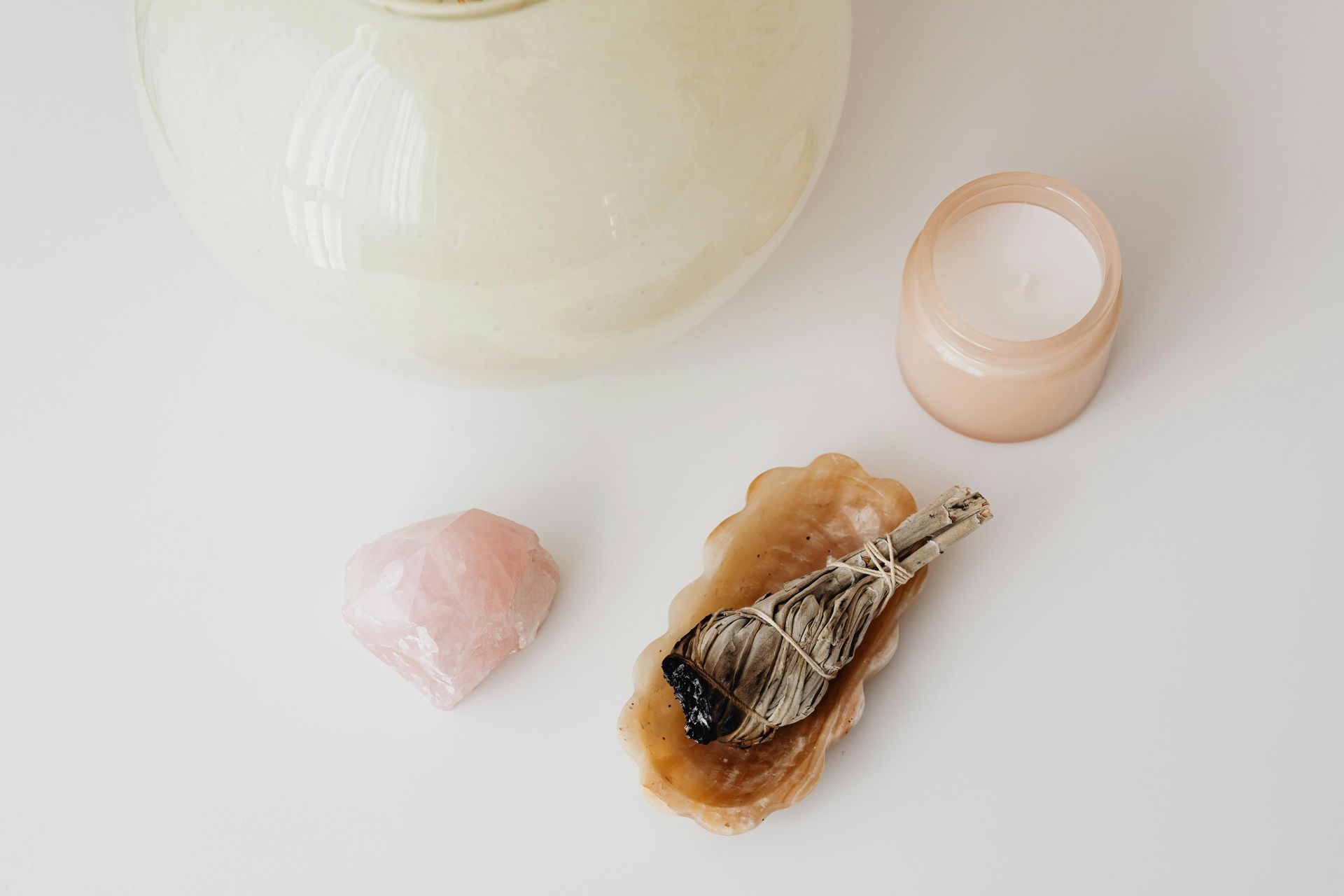 Rose quartz crystal, candle, burning sage, and decorative dish on a white surface.