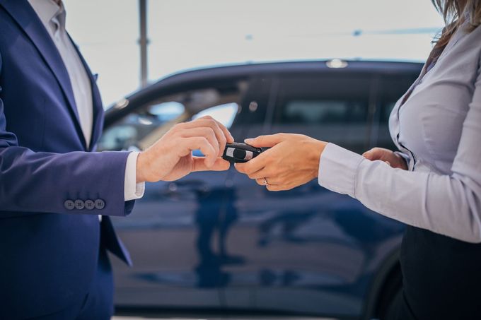 In una concessionaria, una persona in giacca e cravatta consegna le chiavi di un'auto a un'altra, con un'auto blu visibile sullo sfondo.