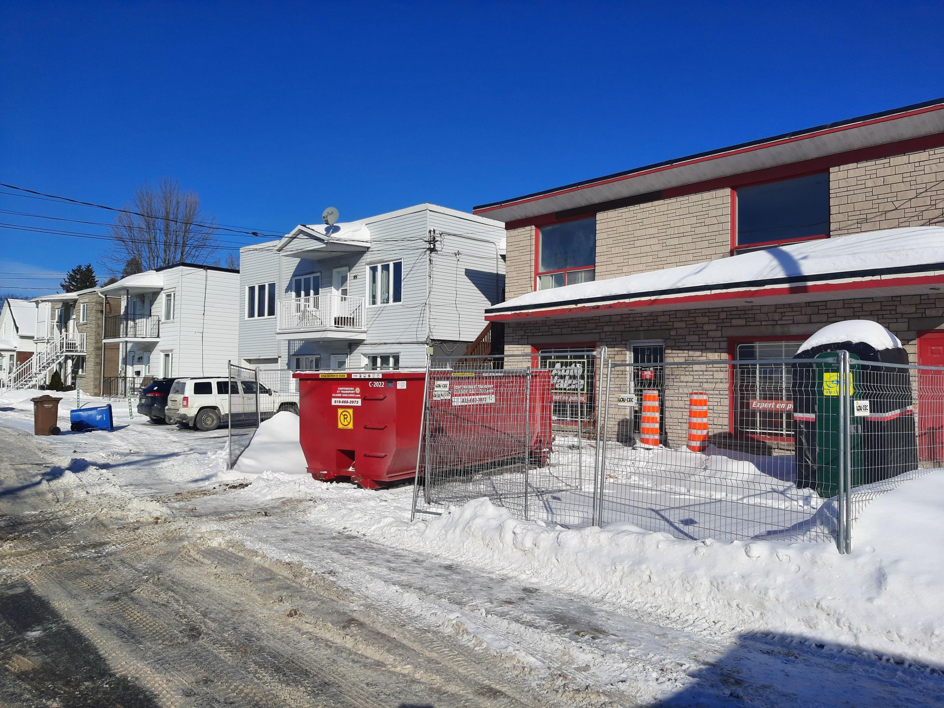 Rue résidentielle enneigée avec des immeubles d'appartements, une benne à ordures rouge et un camion blanc garé sous un ciel bleu clair.