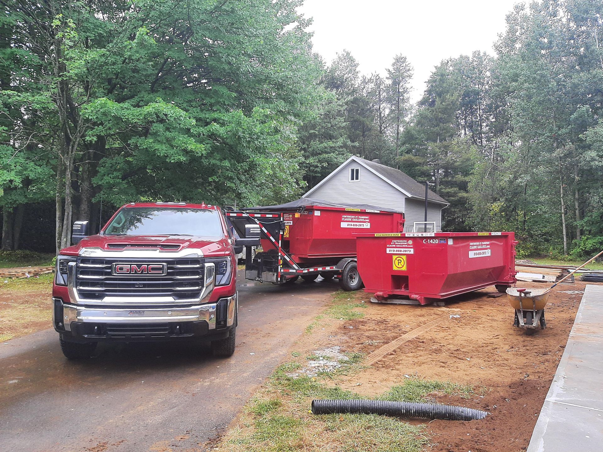 Un pick-up rouge et des bennes à ordures rouges dans une allée à côté d'une petite maison, entourée d'arbres.