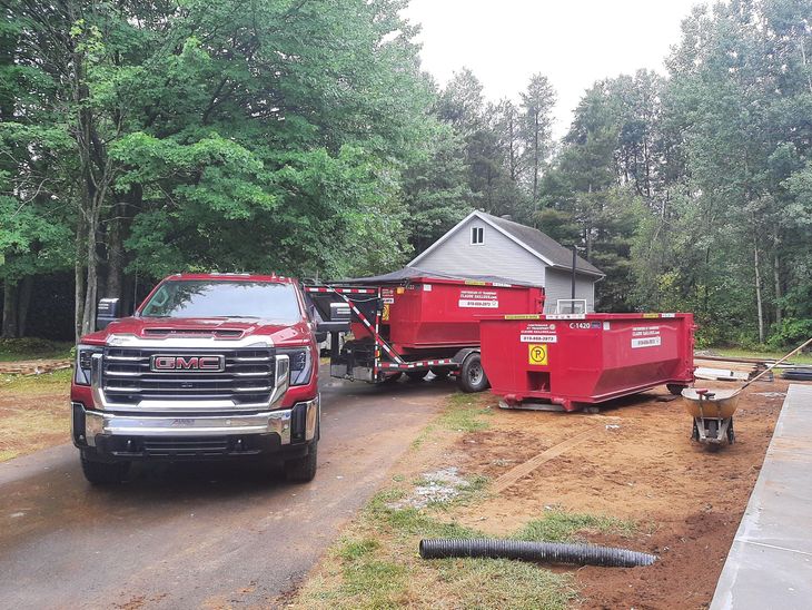 Un pick-up rouge et des bennes à ordures rouges dans une allée à côté d'une petite maison, entourée d'arbres.