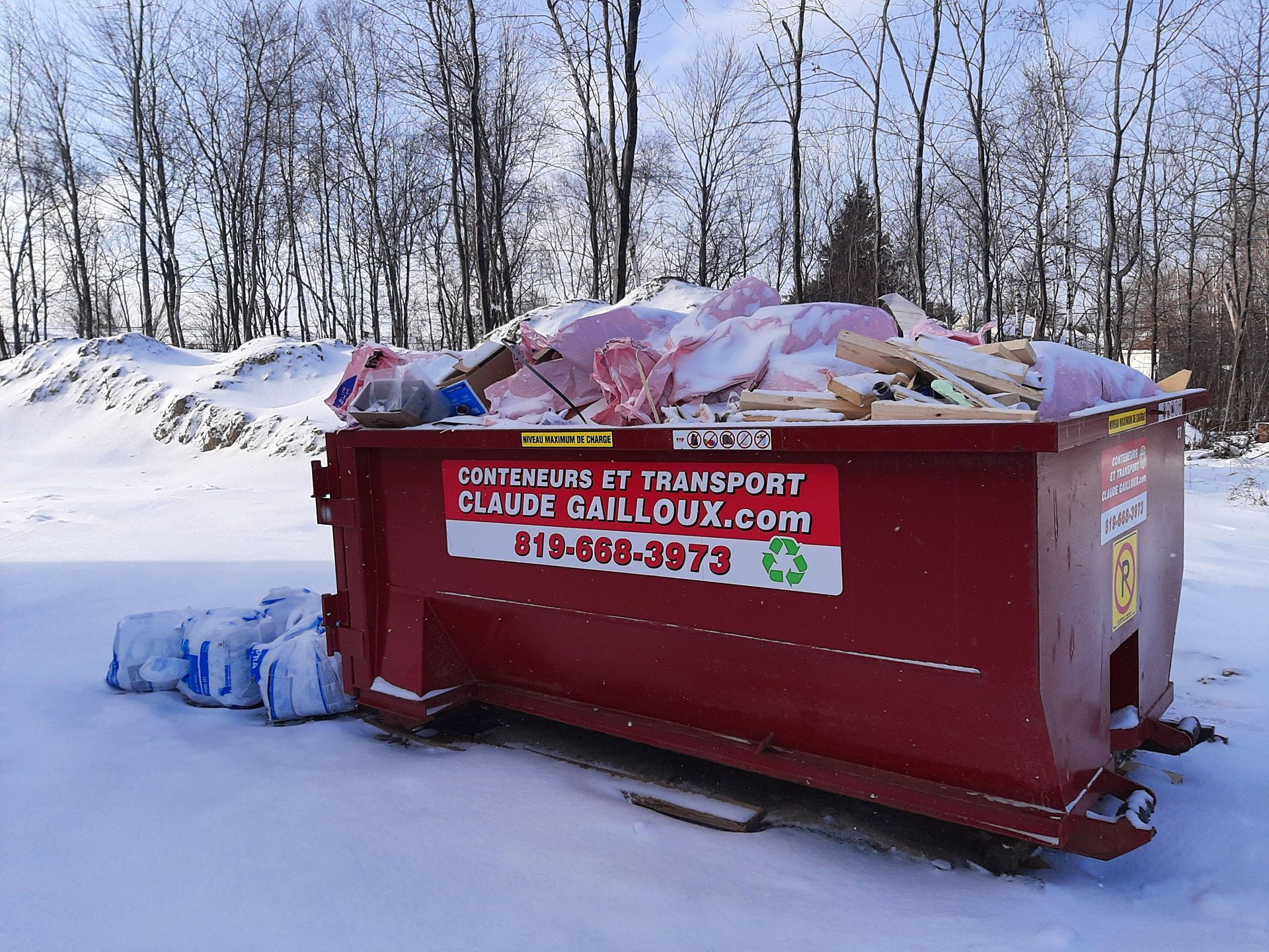 Une benne à ordures rouge débordant de déchets se trouve au bord d'une route enneigée, des sacs-poubelles étant empilés à côté.