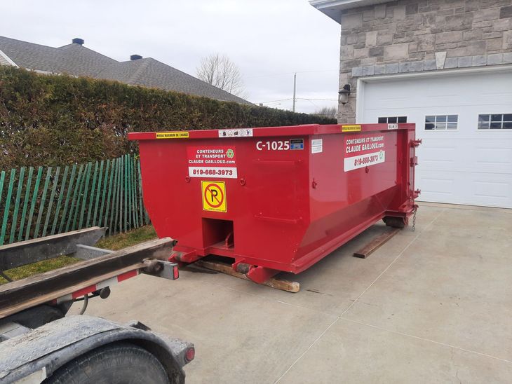 Un conteneur à déchets de chantier rouge est stationné à côté d'un garage, dans une allée.