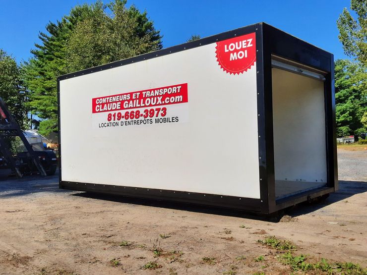 Grand conteneur blanc de déménagement/stockage avec des panneaux rouges, stationné à l'extérieur sur un terrain vague près des arbres.