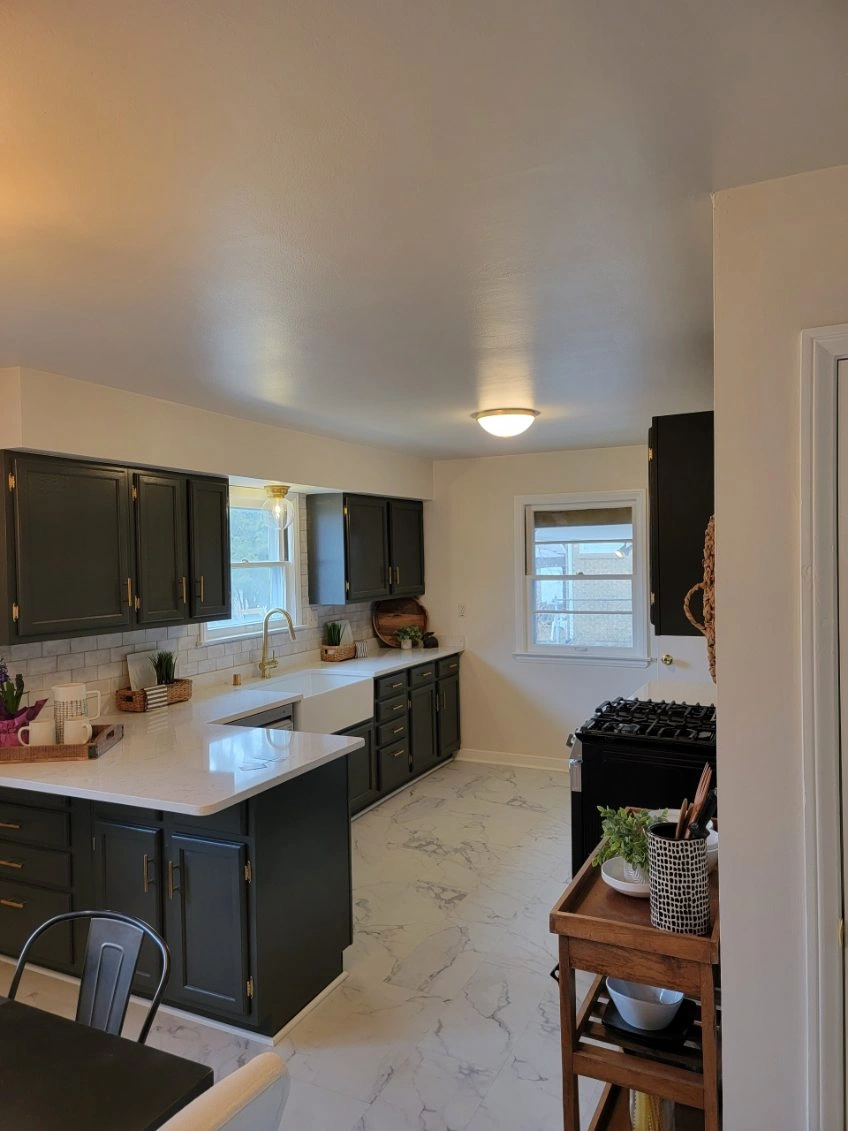 Bright kitchen with dark cabinets, white countertops, and a large window, viewed from the entrance.