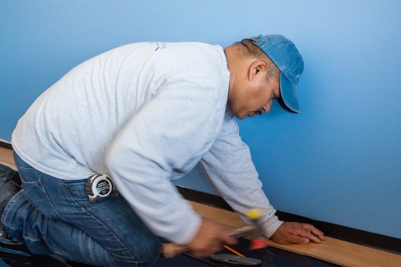 Worker kneeling to install dark flooring, using a tool against a wall in a blue room