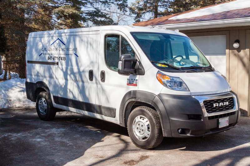 White Amazon delivery van parked in a driveway beside a house, with trees in the background