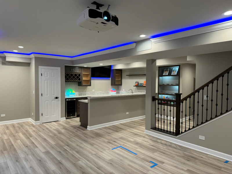 Empty modern basement with gray walls, light wood floors, a bar area, stairs, and blue LED ceiling lighting
