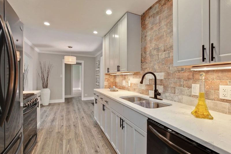 Bright modern galley kitchen with gray cabinets, stone backsplash, stainless appliances, and wood floors