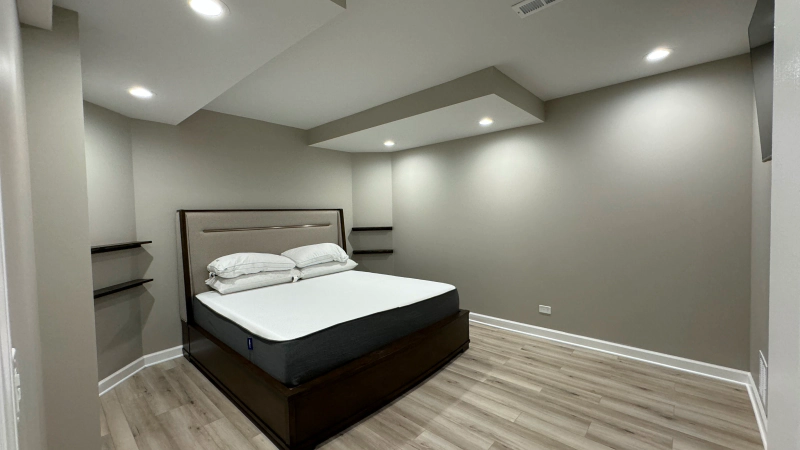 Modern minimalist bedroom with a dark wood bed, gray walls, recessed lights, and light wood flooring.