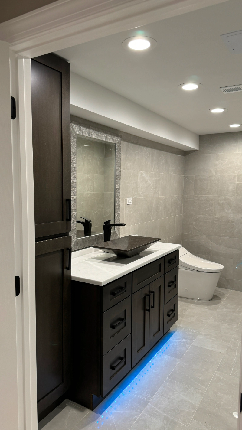 Modern bathroom vanity with lit blue toe-kick, white countertop, large mirror, and toilet in the background