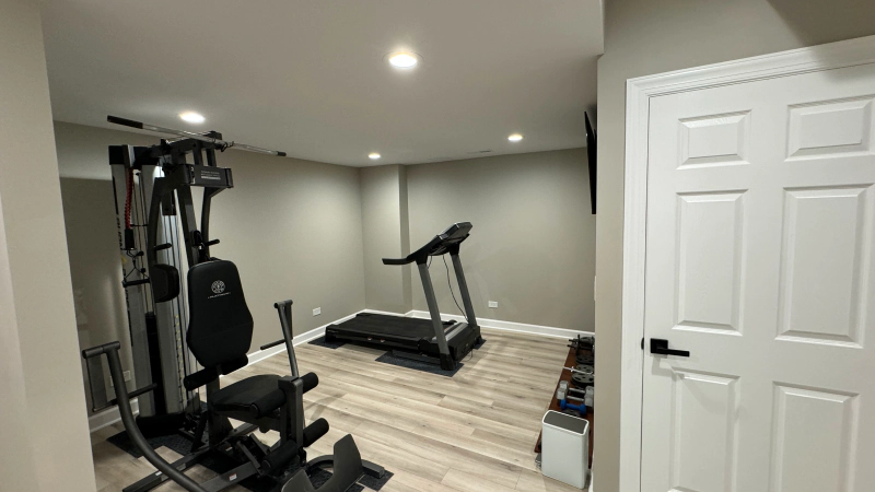 Home gym with treadmill, exercise machine, and rubber flooring in a small room with recessed lights.
