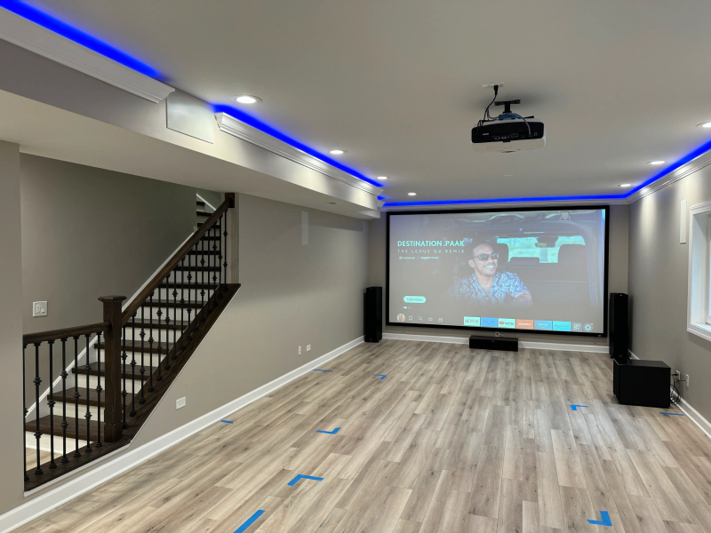 Modern basement home theater with projector screen, ceiling projector, blue LED lighting, and wood-look floors