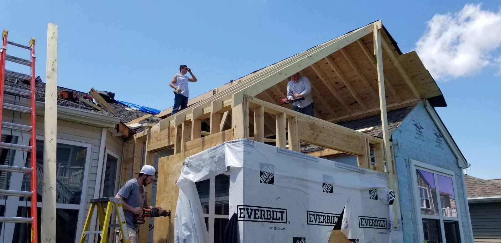 Workers repairing a house roof with exposed framing and white house wrap under a blue sky