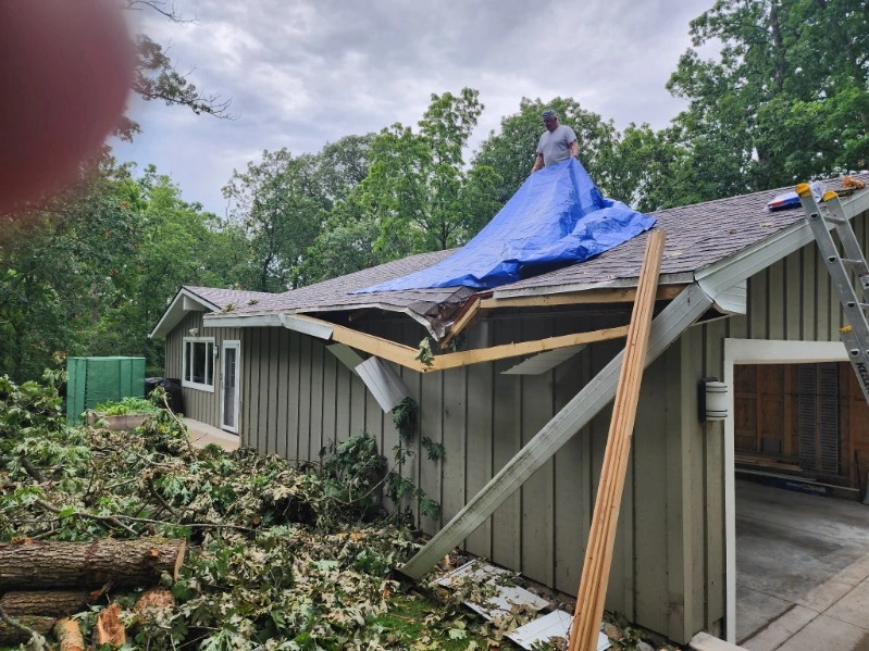 House roof damaged by storm, covered with blue tarp, with fallen debris and a garage visible nearby