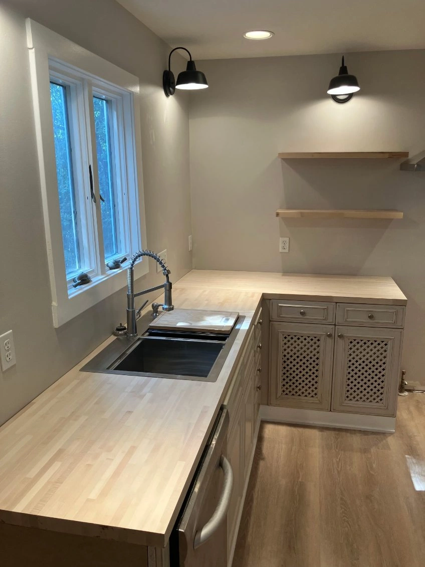 Bright empty kitchen with light wood counters, sink by window, and floating shelves on gray walls