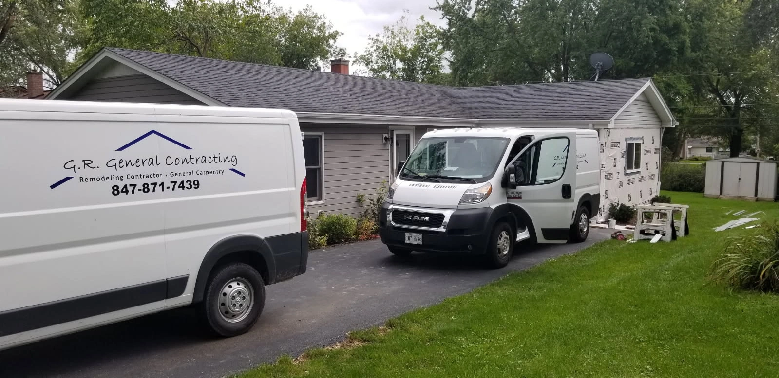 White service van and box truck parked in a driveway beside a single-story house and lawn.