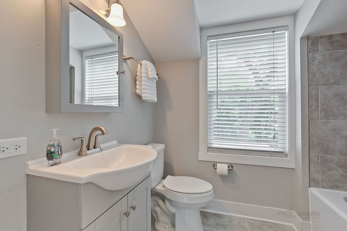 Bright modern bathroom with white vanity, mirror, toilet, window blinds, and tiled shower.