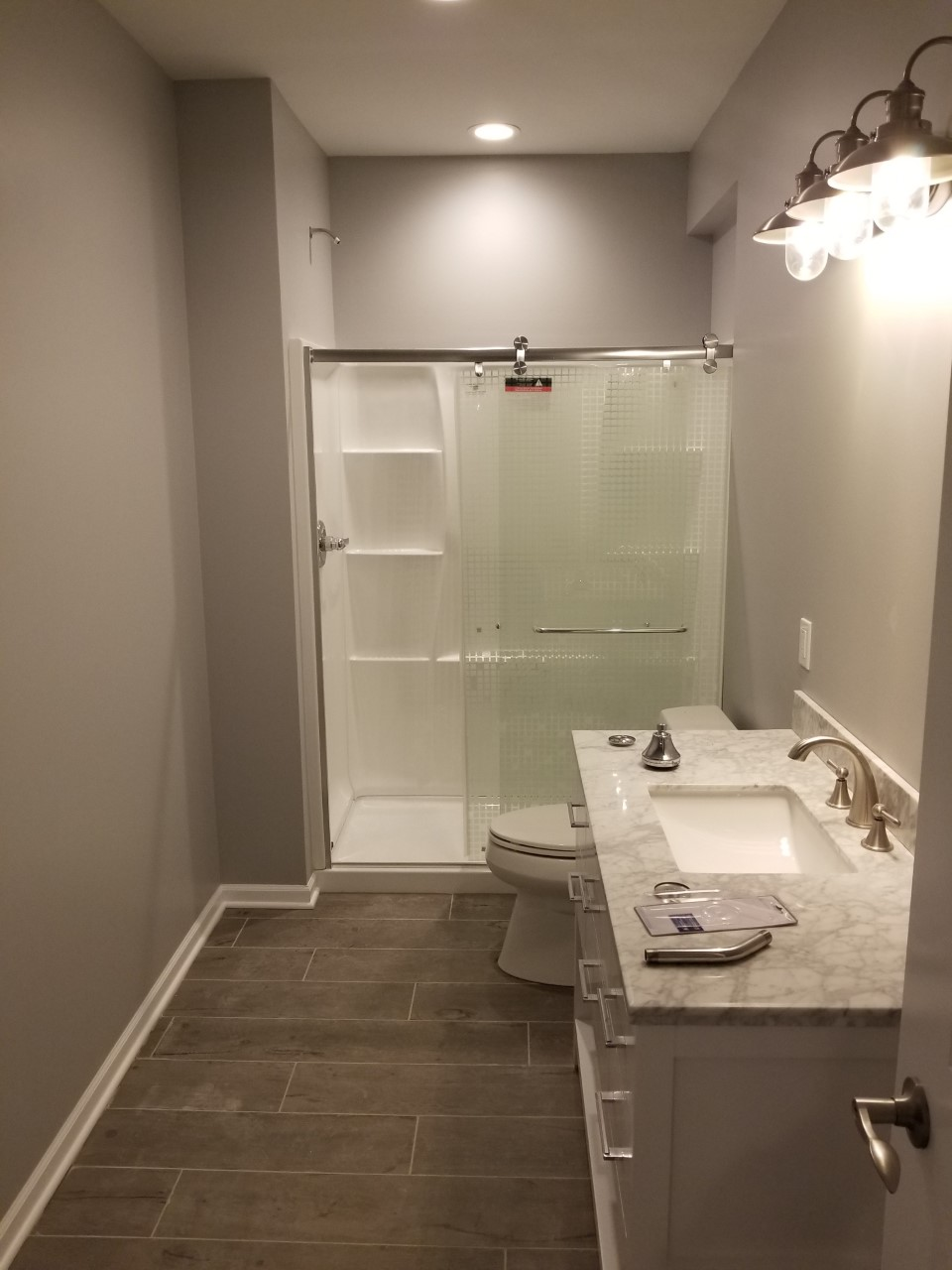 Bathroom with gray walls, dark tile floor, white vanity, shower-tub, and toilet under bright vanity lights