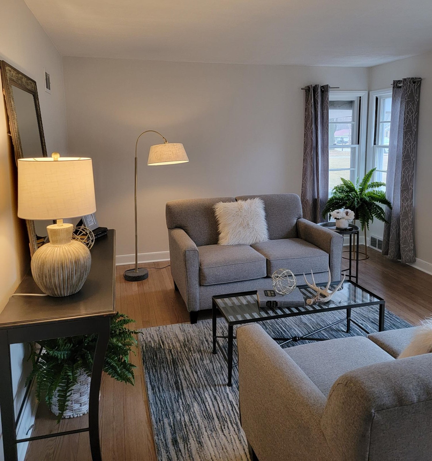 Bright living room with gray sofas, glass coffee table, lamps, rug, and potted plants by the windows