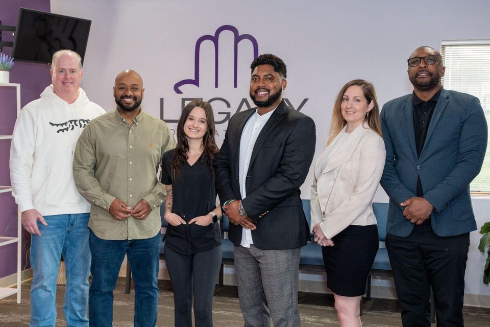 Six people posing indoors in front of a purple wall logo, smiling and holding an award.