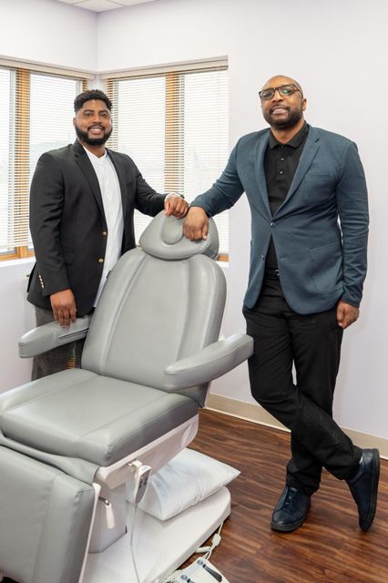 Two men in blazers stand beside a gray reclining chair in a modern office.