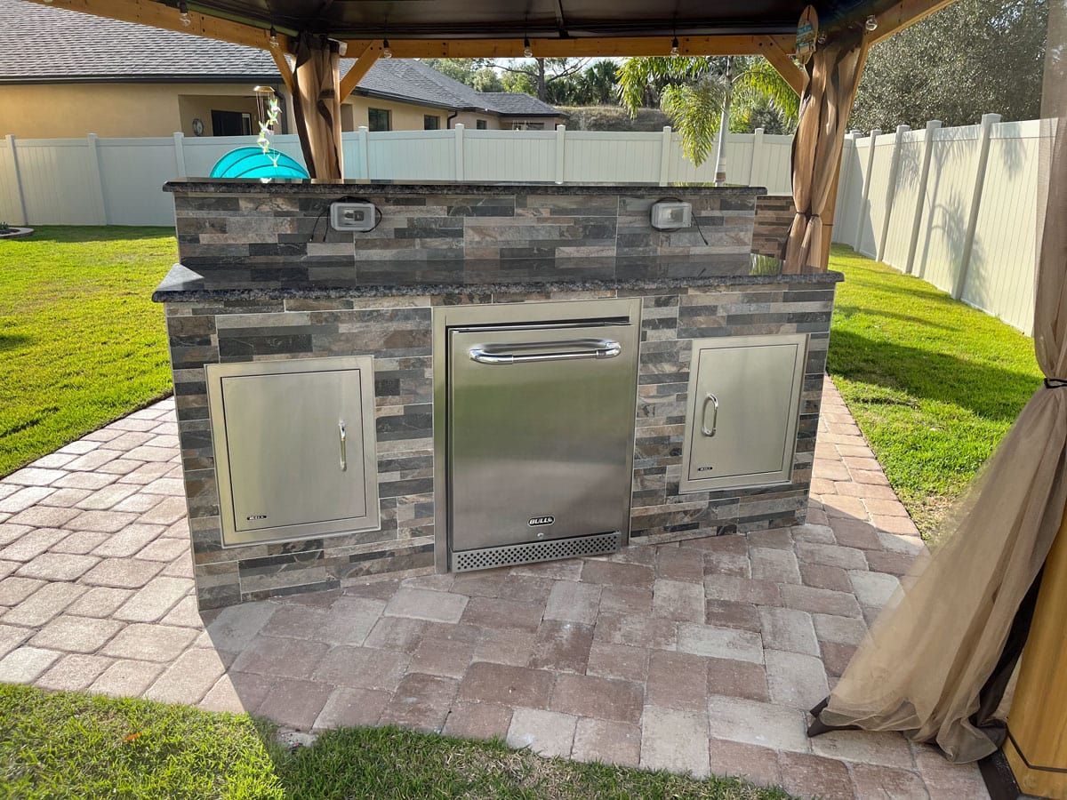 A brick outdoor kitchen with a stainless steel refrigerator under a gazebo in a backyard.