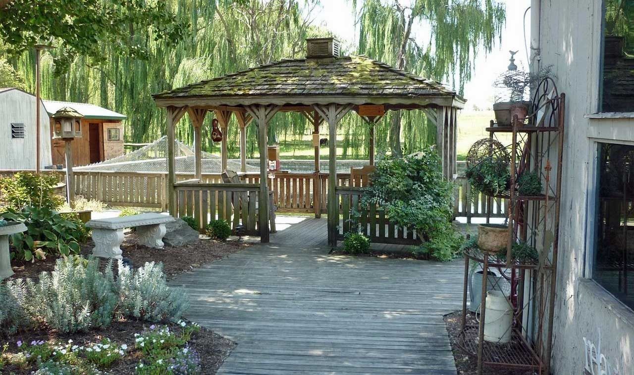 Wooden gazebo on a wooden deck with a garden. Green foliage and trees surround the structure.
