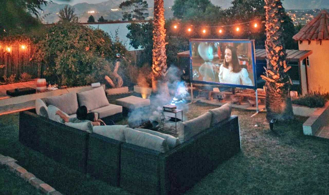 Outdoor movie night: people watching a movie on a screen in a backyard with a fire pit, illuminated by string lights.