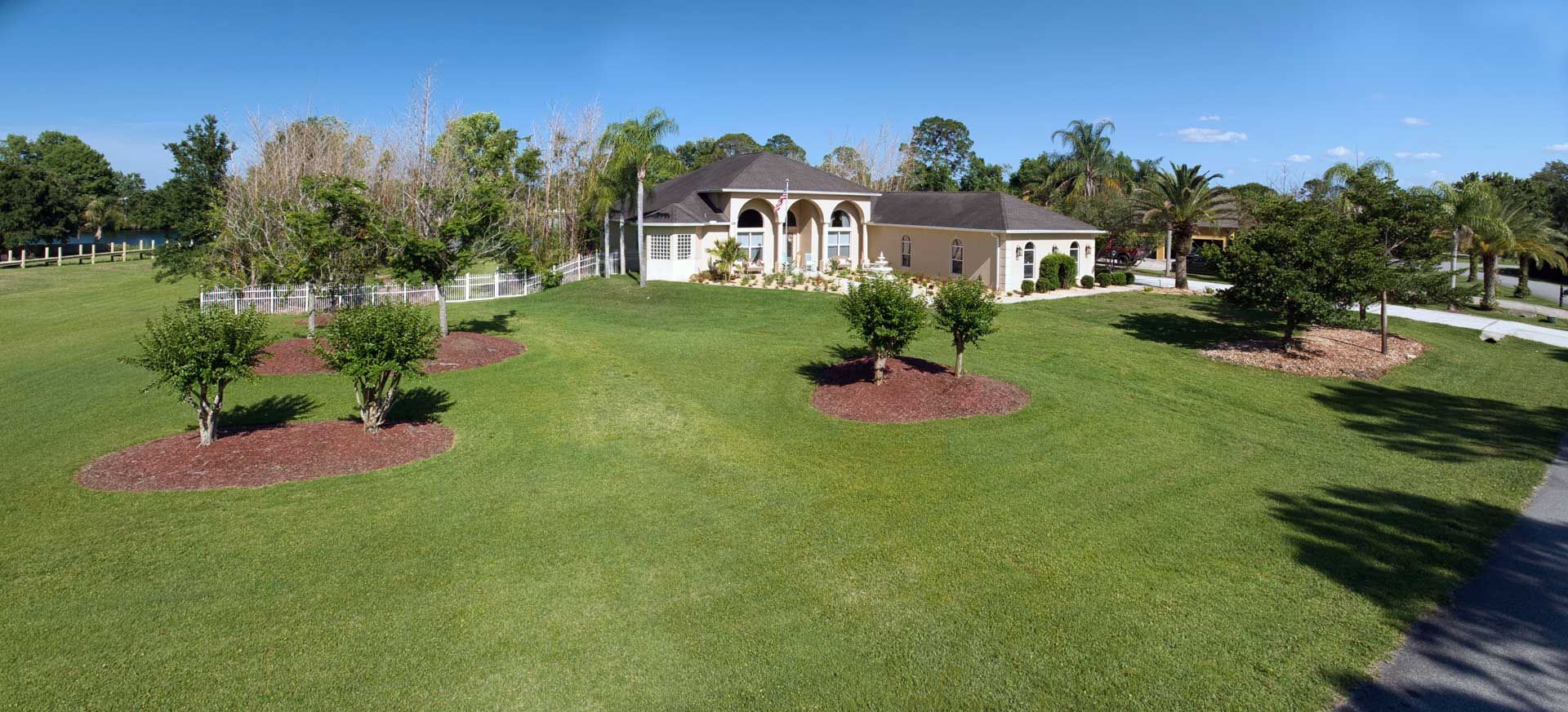 Large single-story house with arched entry sits on a spacious, well-kept lawn with several trees and clear blue sky.