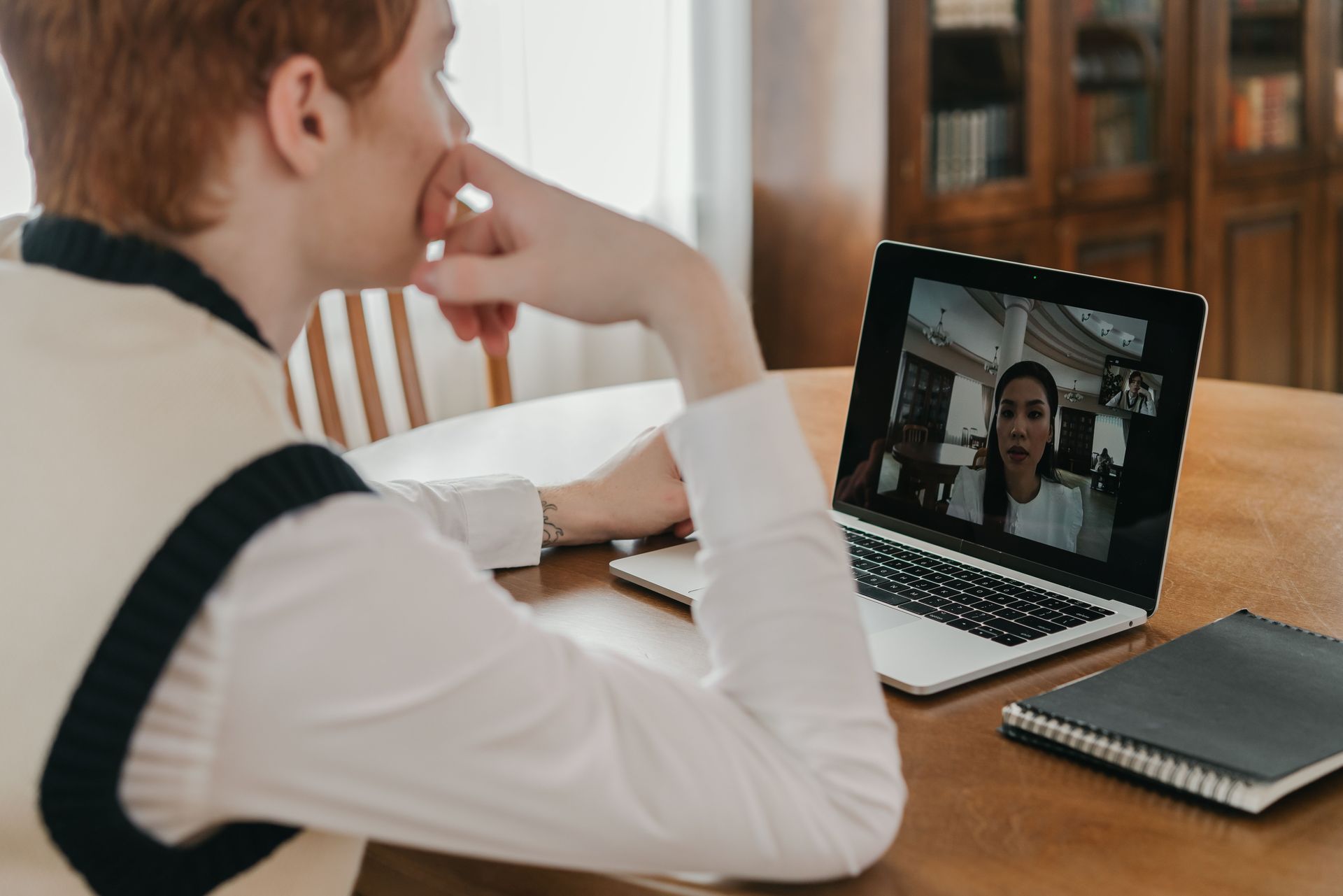 Persona viendo una videollamada en un portátil sobre una mesa de madera, con un cuaderno al lado.