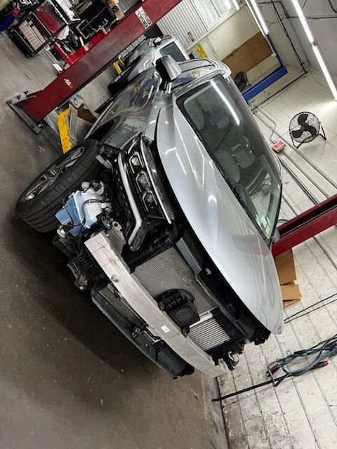 White sports car on a lift in an auto repair shop, front bumper removed and engine exposed