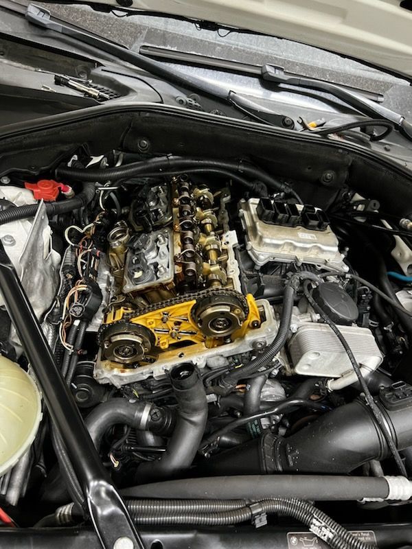 Car engine with valve cover removed, exposing camshafts and timing components in a workshop.