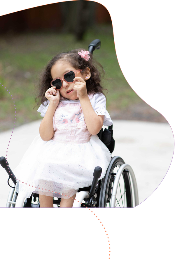 Young child in sunglasses sitting in a wheelchair, smiling outdoors in a white dress