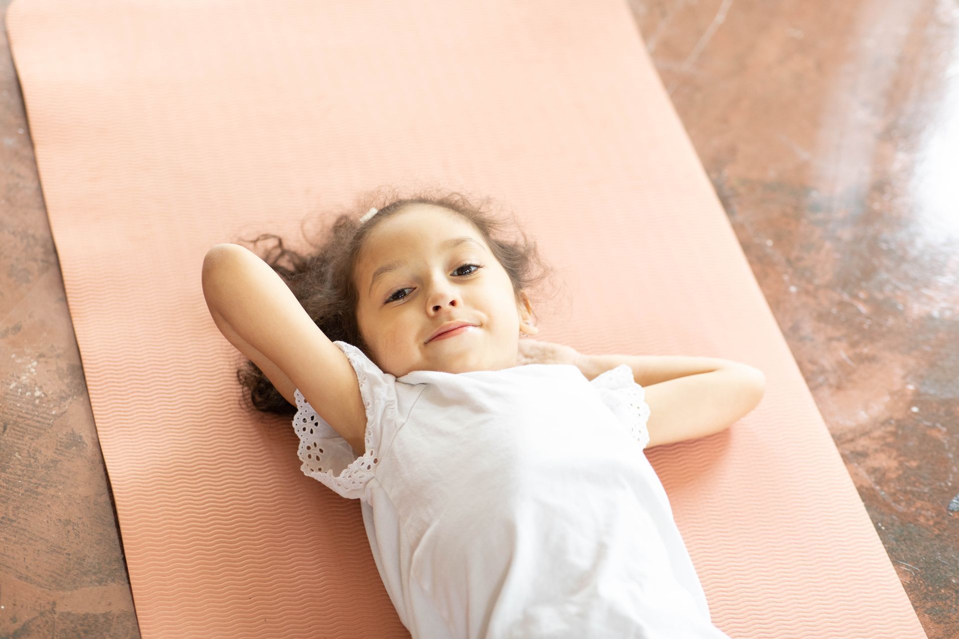 Child lying on a pink mat with hands behind head, wearing a white dress and looking at the camera