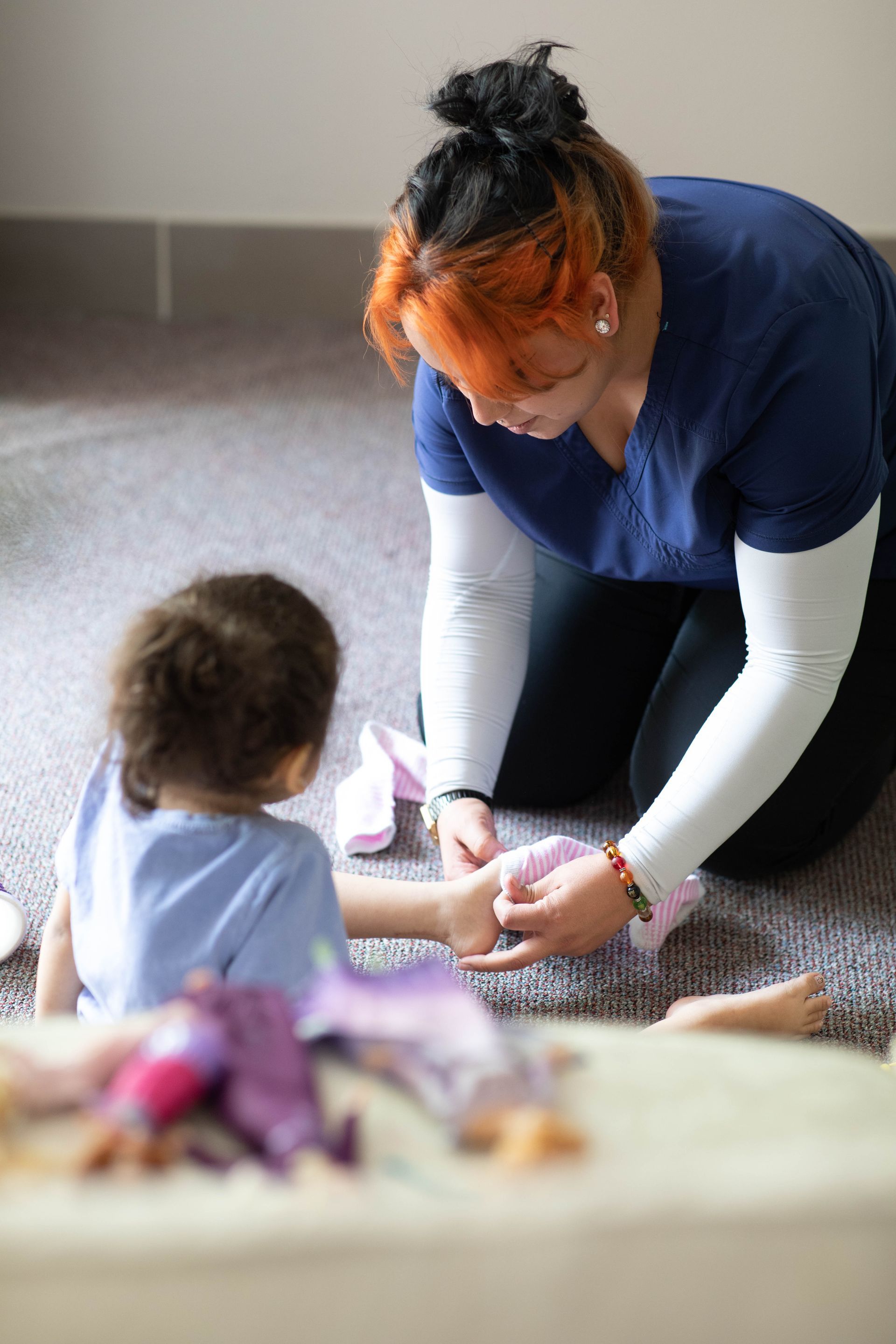 Adult kneels on carpet helping a child play with toys indoors