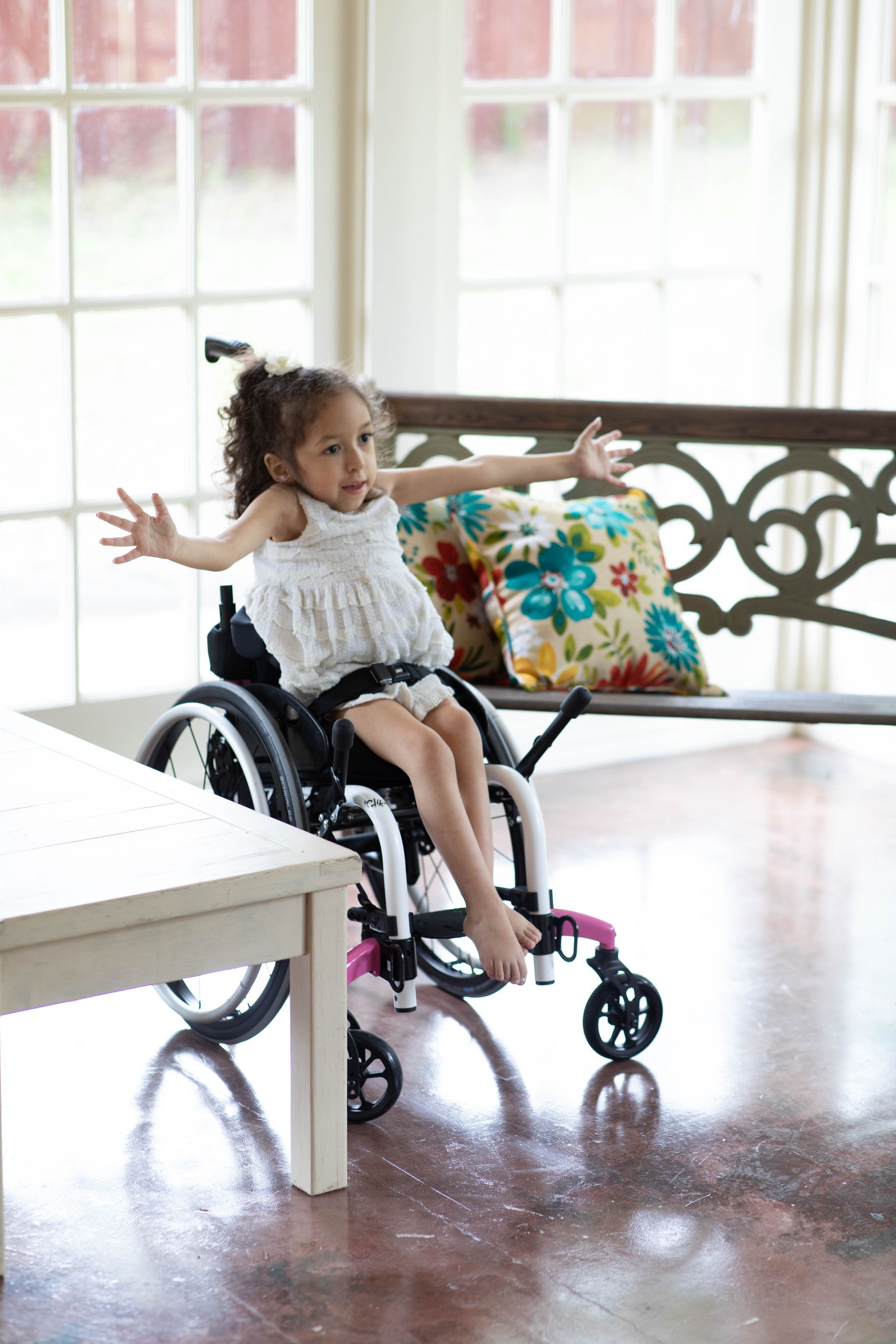 Child in a wheelchair with arms outstretched in a bright room by a table and window.